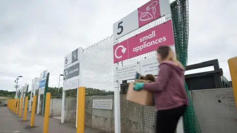 Oxfordshire County Council A tip in Oxfordshire. A woman in a red top and blue gloves is getting ready to get rid of her waste.