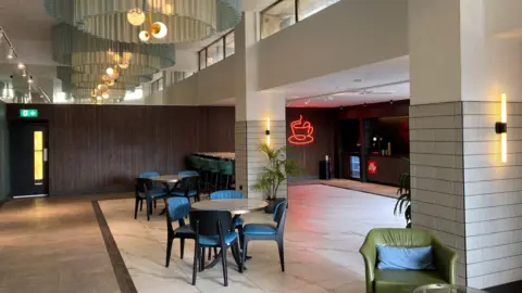 Large curtained ceiling lights with tables and chairs underneath and a red neon coffee cup sign on a wooden panelled wall.