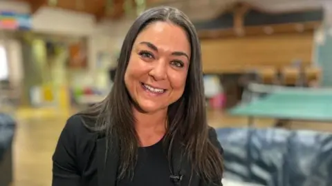 Martin Giles/BBC A woman is sitting in a large room and you can see sofas and a table tennis table in the background. she is wearing a black top and jacket. She is smiling at the camera