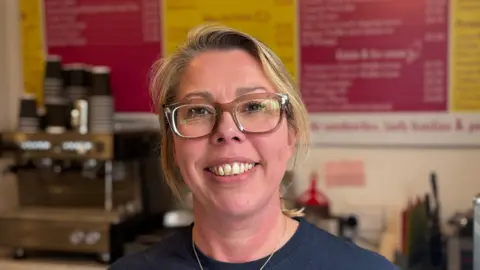George King/BBC A head and shoulders image of Hannah Barton. She is wearing a navy top and glasses and smiling while looking into the camera. 