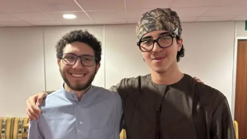 BBC Marwan Ahmed and Mahmoud Elfishawi, two young men standing side by side. Marwan is wearing spectacles, a brown shirt and a patterned bandana around his head, and with his arm around Mahmoud's shoulder. He has dark hair and a close-cropped beard and is wearing a light grey shirt. He is smiling. 