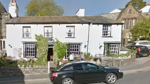 A general view of the outside of Laurel Cottage in Bowness. It is painted white with black window frames and has ivy around its open door. There is a couple sitting at a table outside. 