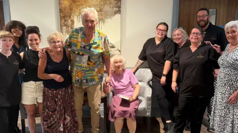 A group of people are stood next to each other with a lady wearing pink sat down in the middle. They are all looking towards the camera smiling. 