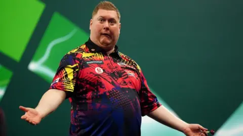 PA Media Ricky has his arms outstretched on stage, wearing a red, yellow and navy blue polo shirt. He is pulling a comical expression.