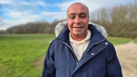 David Burton-Sampson wears a navy blue jacket with grey fur trim around the collar, with a light grey hoodie underneath. He has close shaved hair and is smiling at the camera. He stands in front of an area of grass and trees with blue skies above