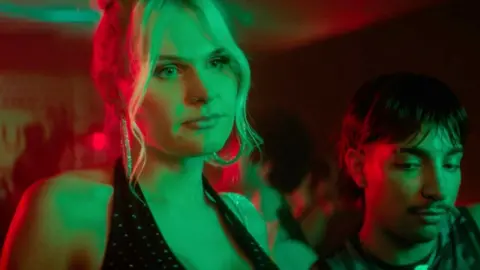 Sasha is wearing a black top. She is in a bar. Green and red lights are shining on her.