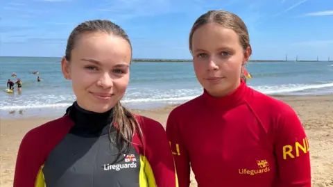 Qays Najm/BBC Phoebe Claybourne and Emily both standing next to each other on the beach at Sea Palling in Norfolk. They are both looking directly at the camera and are smiling. Behind them is the sea and a blue sky.