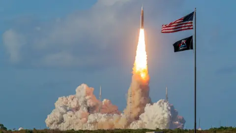 A rocket blasts into space, leaving behind a trail of smoke and fire. An American flag in the foreground.