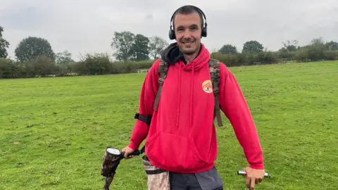 A man in a red top, wearing headphones and holding a metal detector in one hand. He is smiling at the camera.