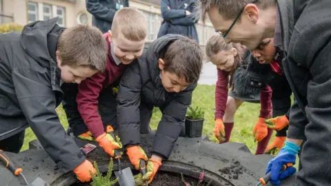 A group of schoolchildren with orange gardening gloves and gardening tools are gathered around a tyre repurposed as a planting area
