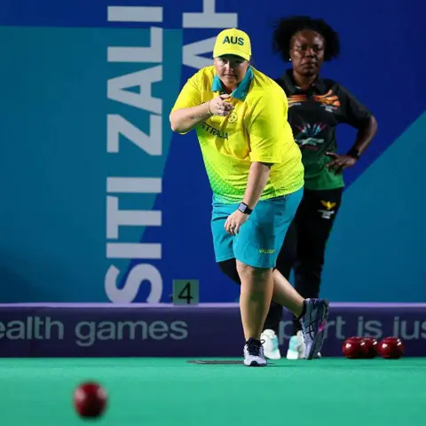 Getty Images Dawn Hayman, a bowler, looking at her shot while standing on an indoor rink. She is wearing a yellow shirt and green shorts. 