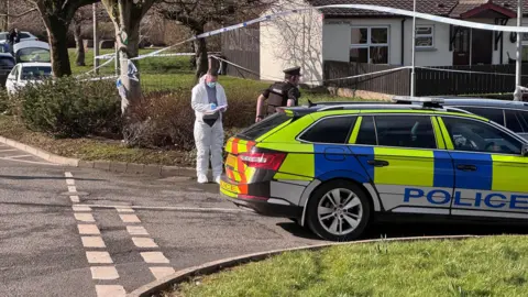 A police car parked on a residential street. Behind the car a man in police uniform and another wearing a forensic suit. Behind them a house surrounded by police tape that is tied to trees and lampposts.