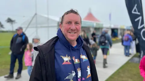 56-year-old Sean Haggerty photographed outside on the maes grounds. He wears a navy hoodie with guinea pigs doing yoga on it, a black raincoat and he smiles at the camera.