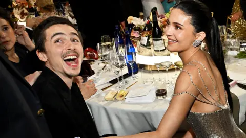 Timothee Chalamet and Kylie Jenner together at the dinner table at the Goilden Globes. He is laughing and she is touching his arm and looking at him