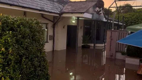 Salique Meah Floodwater surrounds the entrance of a building, with muddy water covering the outdoor seating area and reflecting the doorway, walls and nearby structures. A hedge and patio umbrellas are visible on either side of the flooded area.