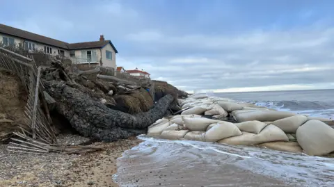 A home on the edge of an eroding cliff in Thorpeness. There is a big cluster of geobag sea defences on the beach in front of it. The tide is washing in.