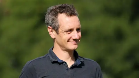 A man with short dark hair smiling and wearing a grey polo shirt.