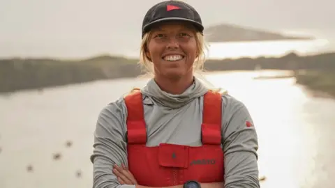 Meg Niblett A woman with blonde hair and a black baseball hat stands looking straight at the camera. She is smiling with her arms folded. She is wearing a long sleeved grey jacket and a red Musto overall.