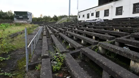 A close-up of the old concrete terraces of Casement, with weeds growing between them. A white building is behind them on the right of the picture and another empty grey two-storey building is on the left.