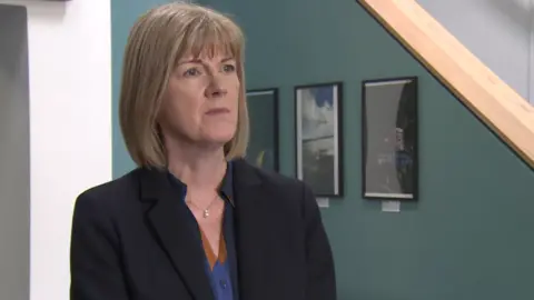 Ms Foster is wearing navy suit with a blue and brown top. She has a blond bob and blue eyes. The walls behind her are blue.