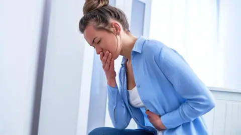 Woman in blue shirt and jeans, clutching stomach with one hand and with one hand over her mouth. She looks nauseous.