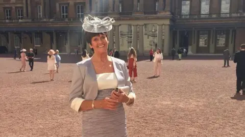 NHS Humber Health Partnership Teresa Jackson standing in the grounds of Buckingham Palace wearing a grey and white dress, with matching hat. Other people and the palace can be seen in the background.