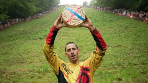 PA Media Chris Anderson stands at the foot of Cooper's Hill holding a wheel of cheese above his head. He wears a yellow and red sports top. 