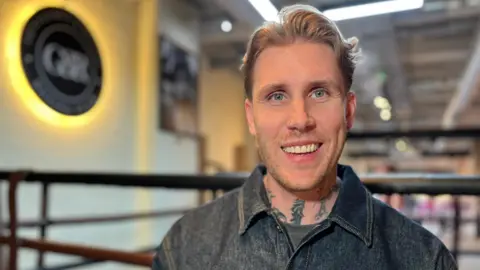 Emma Baugh/BBC A man with blond hair wearing a dark denim jacket smiling in front of a boxing ring. He has tattoos on his neck.