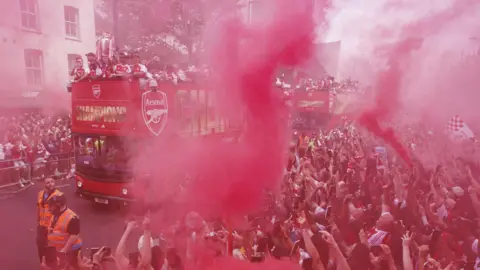 EPA Crowds cheering on the side of a street one of the celebratory open top buses is going through, holding up flares blowing out red smoke