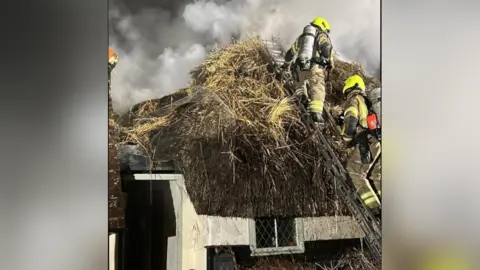 Essex County Fire & Rescue A close-up of a thatched roof, covered in heavy smoke. There are two firefighters on the roof, standing on a ladder. They carry silver tanks strapped to their backs, over their firefighting clothing. They are wearing neon helmets.