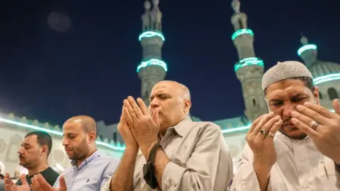 Getty Images Men praying fervently. In the background you can see the mosque which is lit up in blue. It is dark.