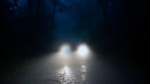 Getty Images A dark wooded road at night, a car is driving towards the camera with bright white headlamps