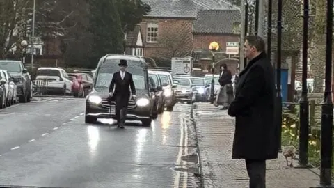 Jenny Kirk/BBC A funeral cortege comes down a street. Some people stand on the pavement wearing black. A funeral director wearing a top hat leads walks in front of a black hearse.
