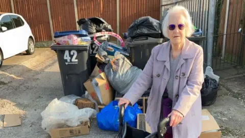 Lorraine Boyce wears sunglasses and a lilac coat and stands near overflowing bins and piles of rubbish in West Heath.