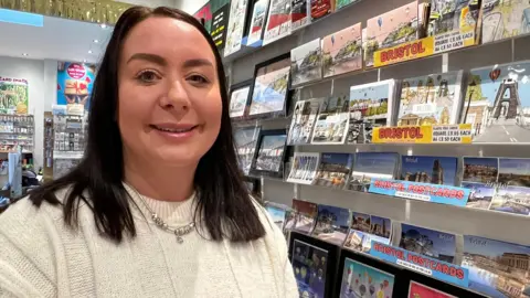 Woman with brown hair and white jumper smiling while stood in front of local postcards inside a shop.