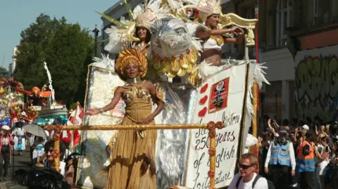 A black woman in a gold dress and golden head-dress dances at the front of a carnival float with the parade stretched out behind her