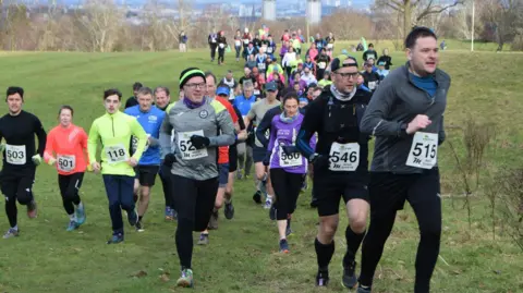 Runners at the Acorn Trails race in Linn Park, Castlemilk, Glasgow on Sunday. There are a number of runners in the image. At the front are three men in dark clothing. A woman in a purple top is behind. They are all mid run. They are in a park with green grass and trees behind them.