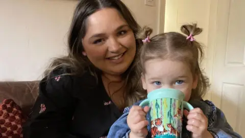 BBC/Cathy Killick A woman with long, dark hair with a child on her knee. The child is holding a blue beaker with tigers on it.