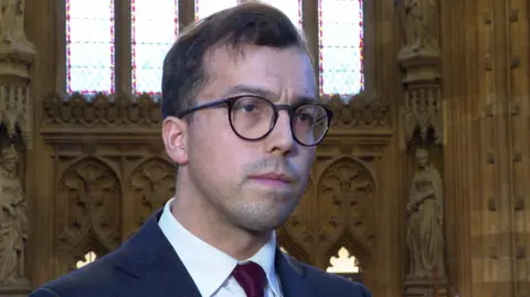 Noah Law MP for St Austell and Newquay stood in Westminster looking away from the camera. He is wearing classes and a blue suit and burgundy tie.