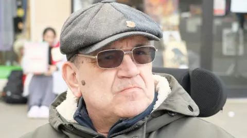 A man in a flat cap with a poppy broach on it, wearing dark glasses looks into the camera. 