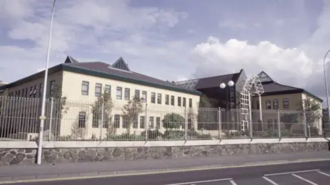 Antrim Crown Court building seen from the other side of the road that runs outside it. Three buildings can be seen behin an iron fence and small stone wall. The entrance to the courthouse is to the right, with a tall lampost to the left.