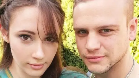 Two people standing close together outdoors, both looking at the camera, with greenery behind them, a woman with long brown hair in a green top, a man with short light‑brown hair wearing a green shirt and a silver chain.