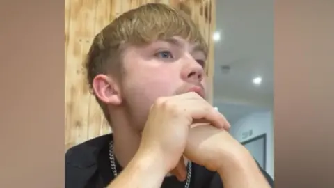 A man with blonde hair, in a black top with a silver chain necklace, looking away from the camera.