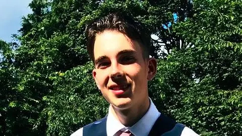 Sam Robinson Photo of a young man, with dark hair, wearing a navy waistcoat, white shirt and pink tie. He is standing in front of some trees in a garden.