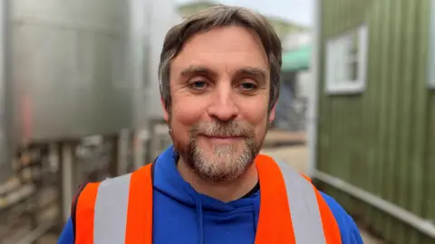 Wye Valley Brewery's managing director Vernon Amor stands outside in front of a green shed and a large steel storage tank. He is wearing a bright blue hoodie and orange hi-vis jacket. 