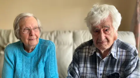 Simon Jones / BBC An elderly couple sat on a white sofa. The lady is wearing a blue jumper and the man is wearing a check shirt. Both look serious. 