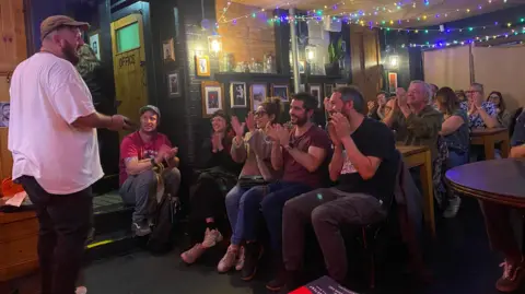 Kev Mud, a comedian in a white t-shirt, performs to a room of people who are applauding him