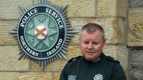 Pacemaker Supt Robert McGowan speaking to reporters at a press conference,  He has short, grey hair and is wearing a green police t-shirt. There is a PSNI logo on the wall behind him. 