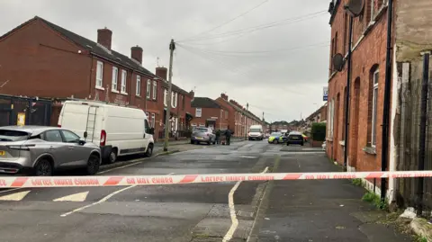 A wide shot of a crime scene in Ainsworth Street in north Belfast. Red police tape is cordoning off a residential street, with cars parked along the footpath. In the distance, a police car is parked opposite a house and officers are outside a car.