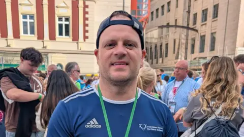 Team NI Richie Sheerin is wearing a navy sports T-shirt and a backwards navy cap. He is also wearing a green lanyard. He is standing in front of a large crowd.